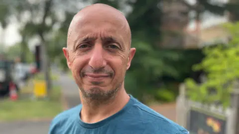 Tom Jackson/BBC A head and shoulders picture of a man with a shaved head and stubble. He is wearing a turquoise T-shirt. He is in a residential street with foliage behind him.