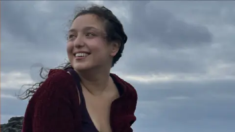 Lara Harris Lara Harris is outside and is looking away from the camera. She is smiling and is wearing a red cardigan. It is cloudy and clearly windy.