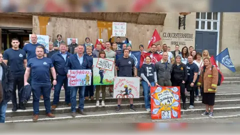 LDRS More than 20 fire service personnel in a mixture of work clothes and non-work clothes, standing in front of Gloucestershire Shire Hall. Some are carrying placards. One reads Firefighters have families too. Another says New shifts will be a logistical juggling act. Another says Our Families matter.