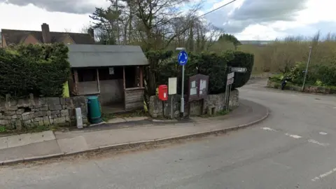 Google A street view image of a small red post box at the side of a road, next to a junction. An old wooden bus shelter is to its left with a notice board and various road signs to the right.