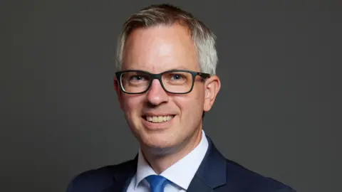 UK Parliament A man with grey hair and glasses wearing a blue suit and a white shirt with a blue tie looks towards the camera, smiling.
