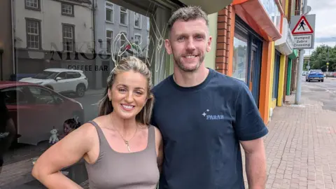 Adrian and Gillian Maxwell stand in front of a row of glass fronted shops. She is on the left and is smiling. She has blond hair and is wearing a grey top. He is wearing a blue tshirt, has short dark hair and is also smiling.