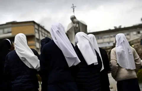 Dylan Martinez/Reuters A group of nuns wearing white veils are seen from behind as they pray in front of a statue of Pope John Paul II 
