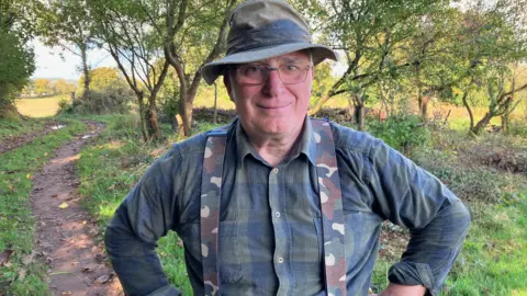 Werner Lothmann, who is wearing a green hat, green shirt and camouflage braces, standing in a green space at Langdon Hospital, Dawlish.