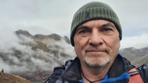 Simon Lucas Simon Lucas, 61, has grey stubble and brown-green eyes. He is wearing a hiking jacket and a green hat and has a cross tied on a brown string around his neck. Behind him are the fells of the Lake District beneath grey skies 