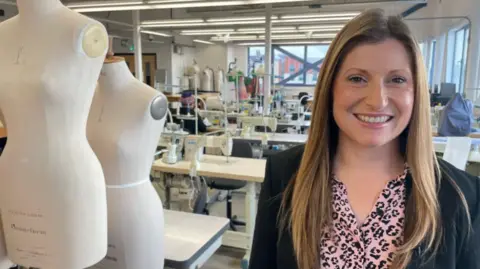 Hannah is pictured in front of a number of tables and artwork. She has blonde hair and is wearing a black blazer.