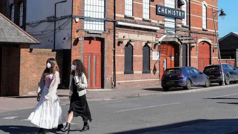 Getty Images Two young people, wearing face masks, one in a white dress one black are walking past Club Chemistry - a red brick building with a black sign saying "chemistry" in white lettering