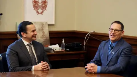 Reuters An image showing Avi Lewis (right) seated at a table opposite from Manitoba premier Wab Kinew. He is wearing a heathered dark blue suit, with a blue shirt and tie, and black-framed glasses. He has short black hair. Wab Kinew is wearing a dark grey suit with a patterned tie, and has long black hair that is tied into a bun. He is seen smiling in Lewis' direction 