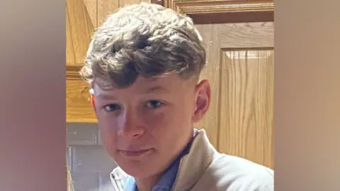 The image is a cropped photo of Connor Magee Campbell. He has short blonde hair and is wearing a blue shirt with a grey jumper.