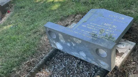 A knocked-over tomb stone bearing the name Caroline Grayson in a grassed cemetery.