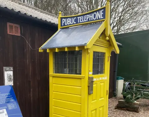 A yellow kiosk, with the words public telephone at the top. Two sides are visible, with both having windows near the top of the kiosk.