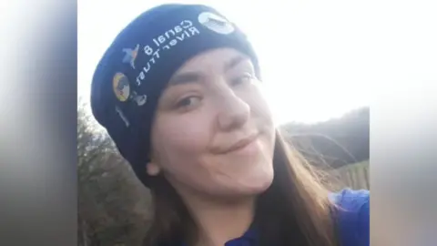 Eve wearing a dark blue hat decorated with multiple colourful embroidered patches. The individual has long brown hair and is dressed in a blue top. The background shows an outdoor setting with trees and a wooden fence.
