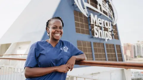 A woman in a blue Mercy Ships uniform stands on the deck of a hospital ship, leaning on a railing, with the charity logo visible behind.