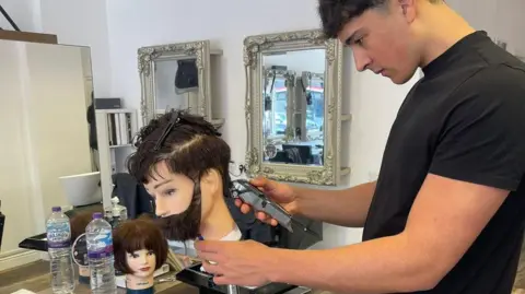 Jude Harvey Foundation Jude Harvey, a young man wearing a black t-shirt, uses clippers to practice cutting hair on a mannequin 