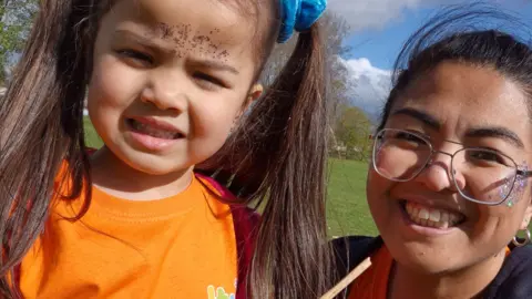 A young girl, with long brown hair in bunches wearing an orange t-shirt, standing in a park next to her smiling mother who is wearing wire framed glasses and has glitter on her face