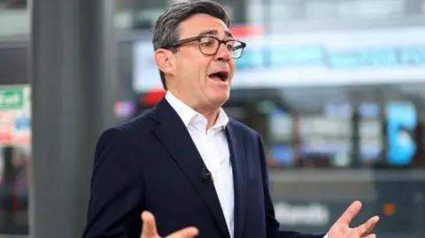 Andy Burnham is wearing a dark-coloured jacket, white shirt and glasses. He has short black and grey hair. He is stood in front of two microphones and 