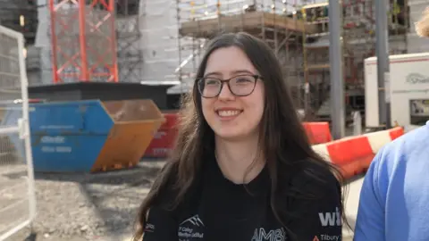 Kayla, with long dark hair and glasses and wearing a black top, standing in front of a construction site