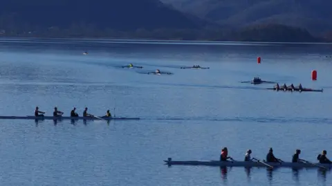 Simon Bamforth Side short of long rowing boats being rowed on the water. There are two orange buoys to the right of the picture. There are mountains in the background. The water is clear. 