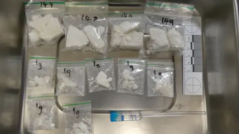 States of Jersey Police Eleven small plastic bags of cocaine laid out on a metal surface. Four of them have "149" written on them and seven have "1g" written on them in black marker pen. There is a paper ruler next to the bags.