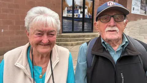 Becca Simmonds/BBC A couple stand in front of a sandstone retail building in a town centre. The woman, on the left, has white hair and is wearing a blue top and cream gilet. He wears a blue baseball cap, pale blue shirt and black gilet, and has a white moustache and blakc-framed glasses.