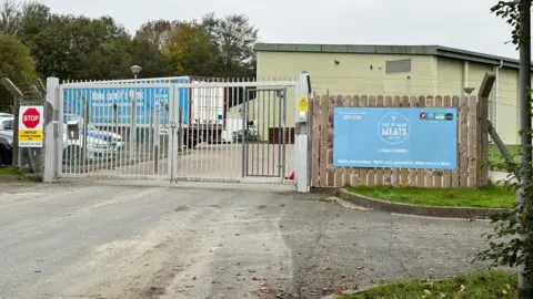 BBC The entrance to the Isle of Man meat processing plant. There is a large metal gate across the entrance and a light blue sign on the right featuring the Isle of Man Meats logo.