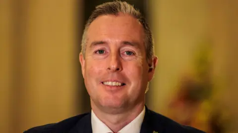 Paul Givan smiling at the camera, he has greyish black hair, he is wearing a black blazer and white shirt