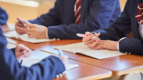 Getty Images Anonymous students wearing uniforms and sitting at desks. The photo is cropped so only the students' arms are visible. They are wearing navy blazers and burgundy ties.