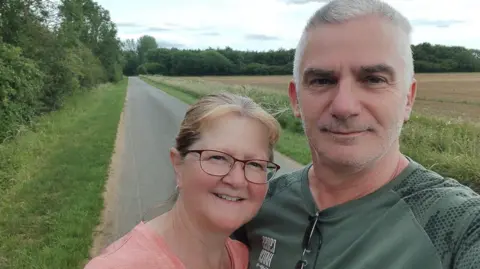 Mike Rickard A man and a woman pictured on a country lane. He has grey hair and is wearing a green top. The woman has light-coloured hair and is wearing glasses and a peach-coloured top. 