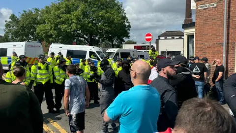 Protesters gathered in Stoke-on-Trent on Saturday, lining up in front of police. One person is in a full face  mask, another with a scarf pulled up to just below his eyes, while others stand watching police