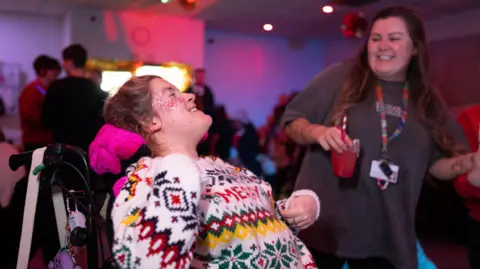 Woman in a fair isle Christmas jumper is dancing. She's a wheelchair user and is smiling. She has glitter on her cheeks. She's dancing with another woman who has long brown hair and is holding a red coloured drink. 