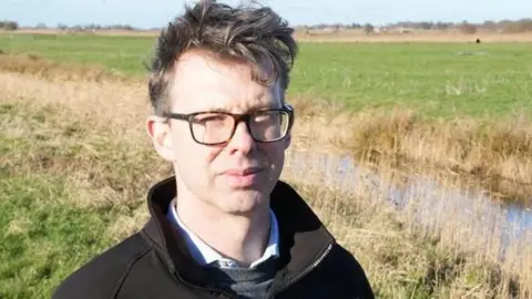 John Fairhall/BBC Eliot Lyne is looking at the camera and is stood in a field. He is wearing glasses and is wearing a black sweatshirt, shirt and grey jumper. 