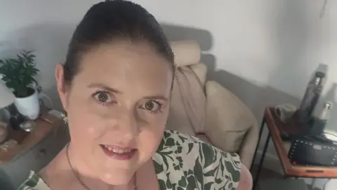 Rachael Johnson who is 41 years old. She is smiling and looking up at the camera. She has hazel/brown eyes and is wearing a short sleeve summer dress which is cream with a khaki leave pattern.