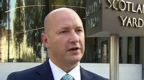 Commander Dominic Murphy is standing outside the Met Police headquarters in London. A sign behind him says "Scotland Yard". He is balding and is wearing a blue suit jacket, a white shirt and a patterned light blue tie