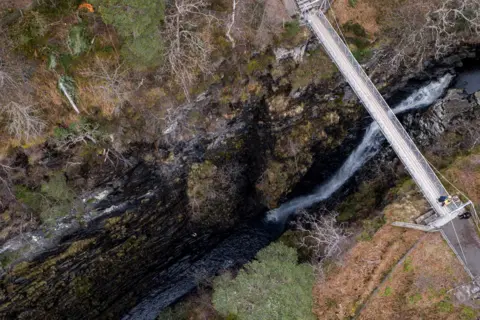 NTS An aerial image of part of Corrieshalloch Gorge. There is a narrow footbridge over the gorge, which has steep sides with water flowing at the bottom of the crags.