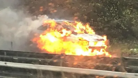 A car is seen from the other side of a central reservation made up of metal barriers. The car is on fire with flames at the lower front end of it, and smoke rising up out the back. It is a small silver car.