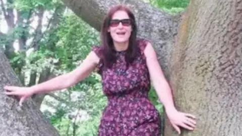 A middle-aged woman with long reddish hair and wearing a floral dress and sunglasses stands smiling between two big tree branches. 