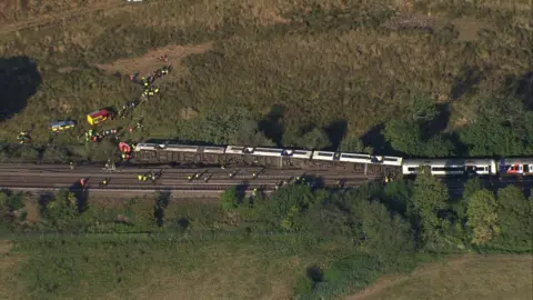 Eddie Mitchell An aerial photograph shows a passenger train with multiple derailed carriages near Lewes in East Sussex. Carriages are off the tracks, with some lying on their side beside the railway. Emergency service vehicles and dozens of responders can be seen on the railway line and in a nearby field surrounded by trees and open countryside.