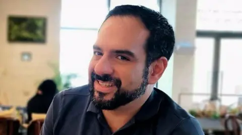 Man with black hair and a beard smiles at camera