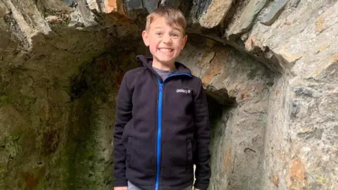 Contributed Cassian pictured outside a stone archway that looks like part of a castle. He wears a navy blue gelert fleece zipped up jumper and grey tshirt. He is kind of smiling or grimacing with his eyebrows raised. He has short fair hair.