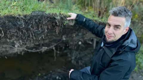 Mr Edwards has short dark and grey hair and a black beard, He is wearing a black coat as he kneels down next to tree roots. His hand is on the grass around the roots as he turns to look at the camera.