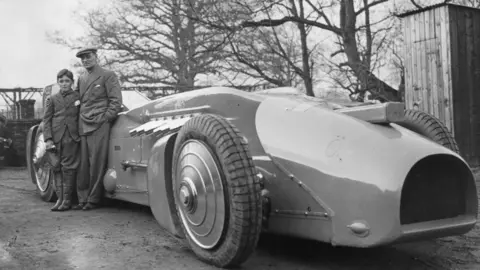 Getty Images A man stands with a smaller boy next to a car with large wheels and a sloped nose. The image is black and white.