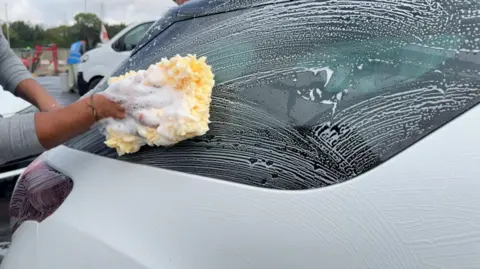 Image shows a white car and black window. It is covered in soap. From the left of the image, there is a hand holding a yellow sponge. He is covering the window in soap. 