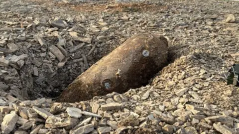 The picture shows a large, rusted cylindrical object, a a German SC250 air-dropped bomb, lying on its side in a shallow pit at what appears to be a construction or excavation site. The object is partially embedded in loose grey rubble made up of broken stone and aggregate.
