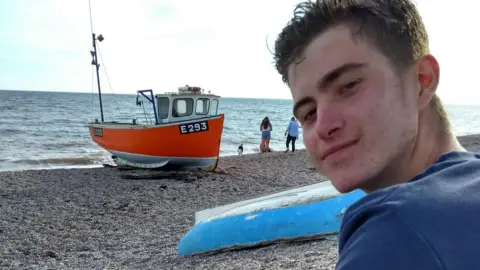 Family handout Gus sits on a beach in swimming shorts and his bare legs and a long sleeve t-shirt and looks over his left shoulder and smiles at the camera