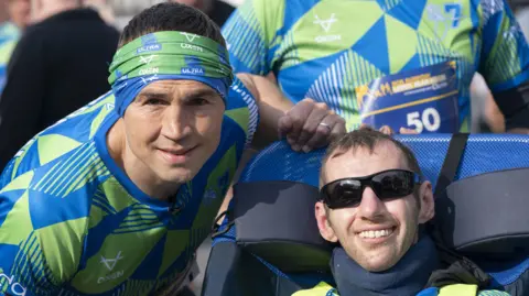 PA Media Kevin Sinfield (left), and Rob Burrow, smiling together at the start line of a race, with Sinfield wearing a green and blue running top and bandana. Rob Burrow is wearing sunglasses and in a wheelchair.