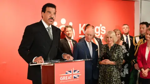WireImage / Getty Images Lionel Richie speaks at a podium with the UK flag on it that reads 'Great Britain and Northern Ireland', with the King and Queen and other guests in the background, at a King's Charity event in New York on Wednesday.