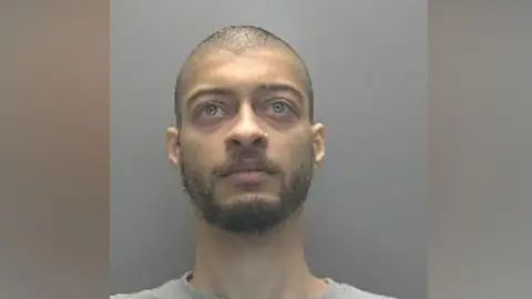 A police custody photo of Liam Rush, with tightly cropped brown hair and a beard. He is wearing a grey T-Shirt and is standing in front of a grey wall. He is looking above the camera.