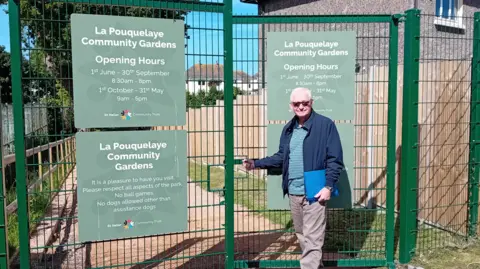 St Helier Community Trust Vice Chair of the St Helier Community Trust stands in front of green metal gates into the community garden which lead down a path with a fence and houses on one side.