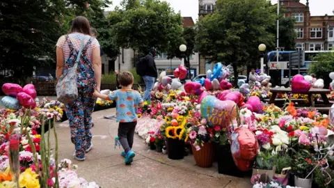 Reuters A mum, in flower-patterned dungarees and a striped T-shirt, and her young son, in a blue and orange T-shirt and black trousers, walk past a large number of bouquets, balloons and plants left in tribute to those killed in the Southport attack