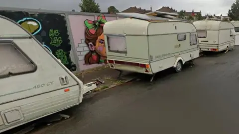 Google A row of white caravans on a city street in an industrial area of Bristol. Brightly coloured murals are visible on building site hoardings in the background. 